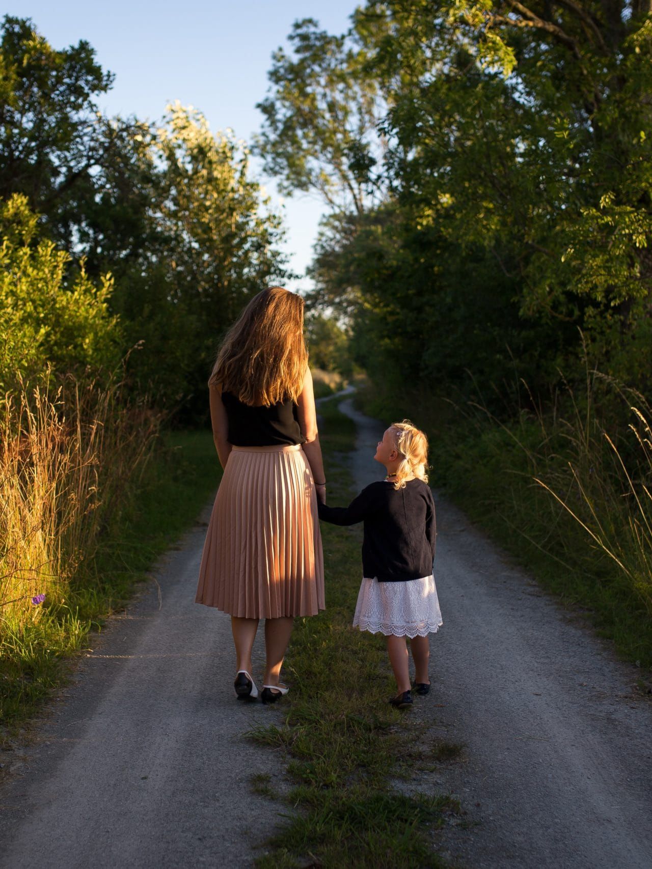 woman and girl walking on road surrounded by green grass