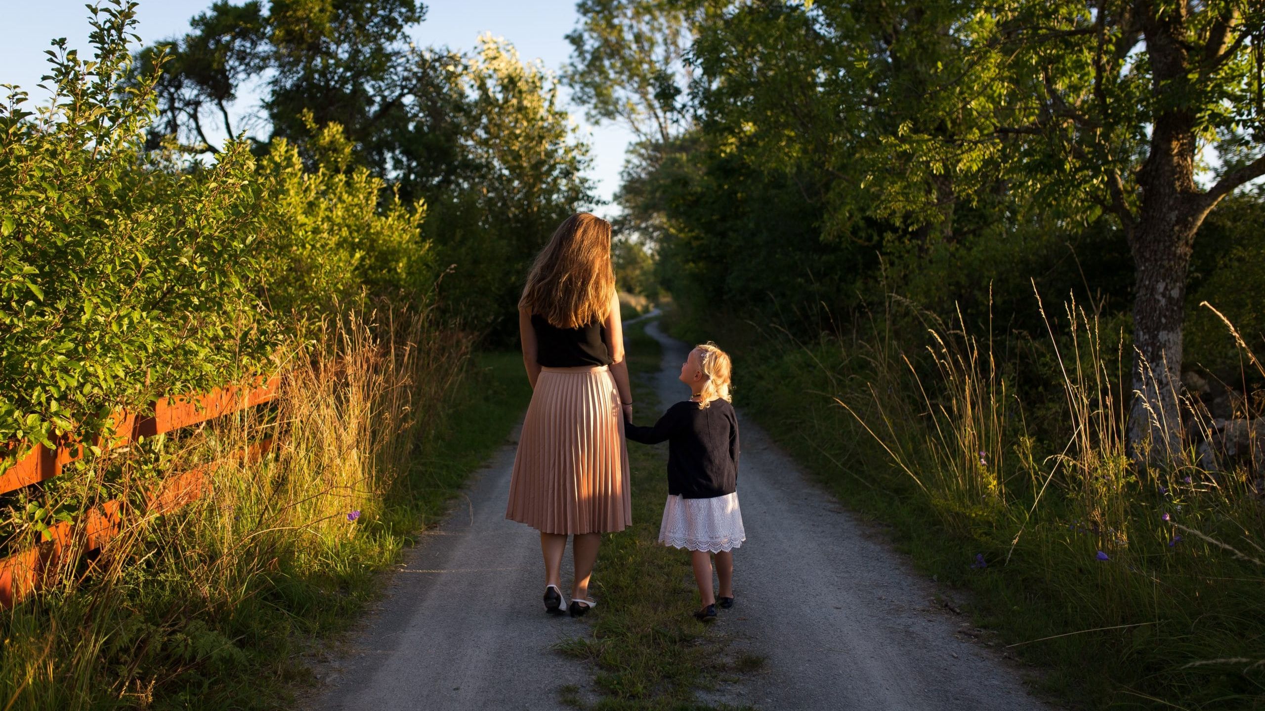 woman and girl walking on road surrounded by green grass