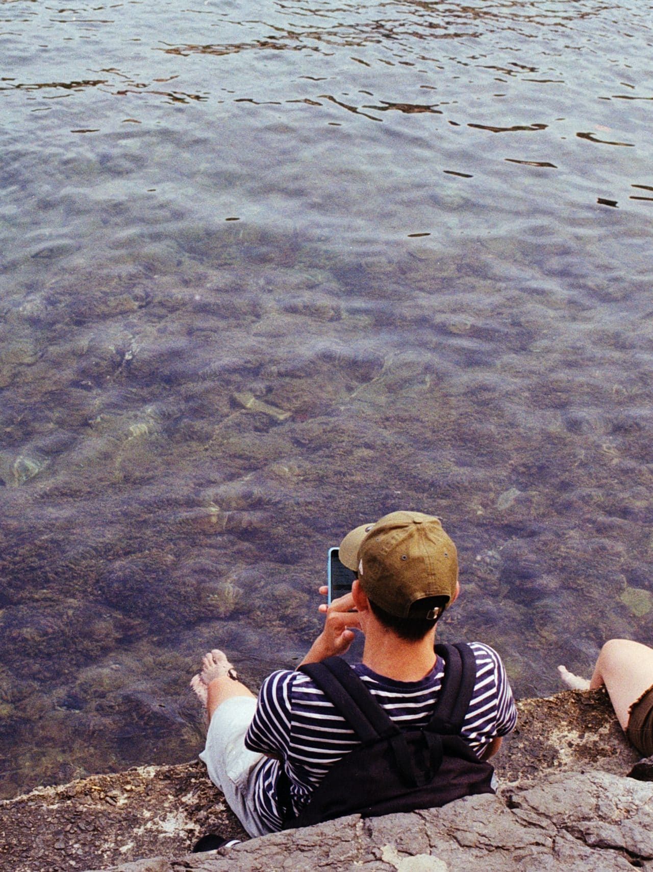 man in black and white stripe shirt sitting on rock near body of water during daytime