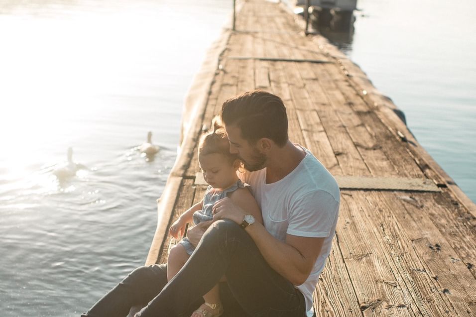 man and girl sitting on brown dock near boat and two white ducks during daytime